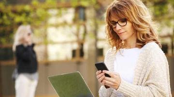 Image of a person sitting on a park bench looking at her tablet and laptop.